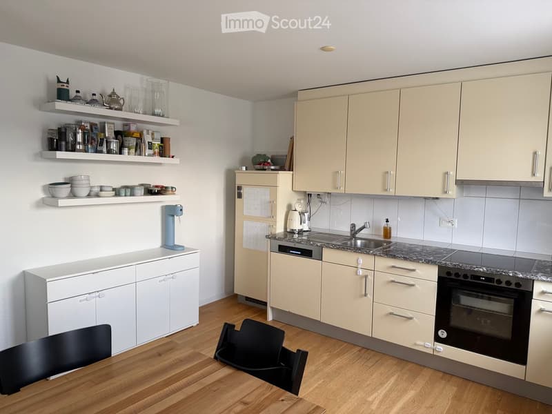 Modern kitchen with light-colored cabinets, granite countertop, and various appliances including an oven, coffee maker, and sink. Shelves hold dishes, cups, and other kitchen items. A small dining area with a table and chairs is visible.