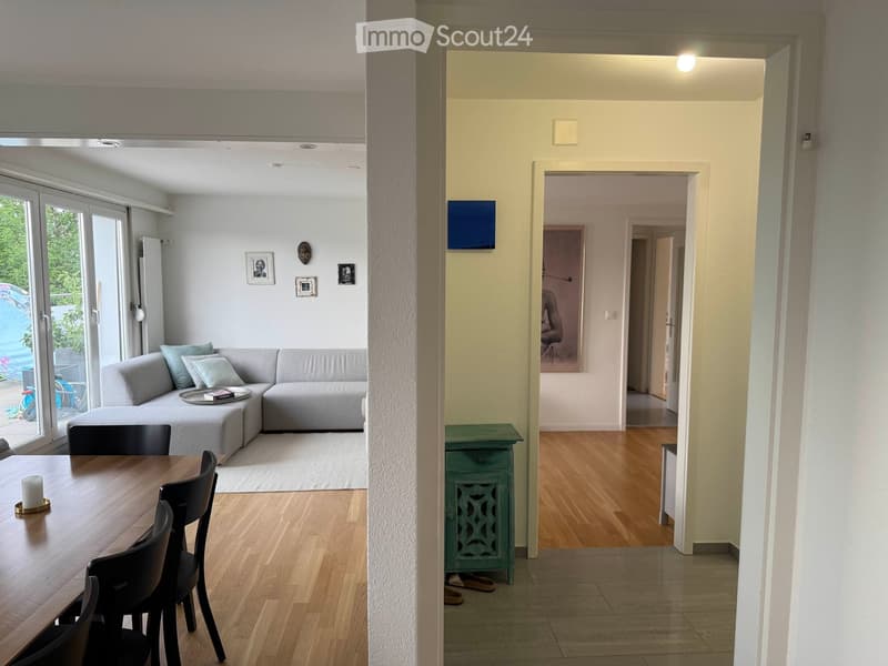 Open-plan living and dining area with a grey sectional sofa, wooden dining table with chairs, and a small green side table near the entrance.