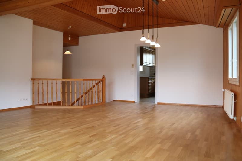 Empty room with wooden flooring, white walls, wooden staircase railing, and visible kitchen area in the background.