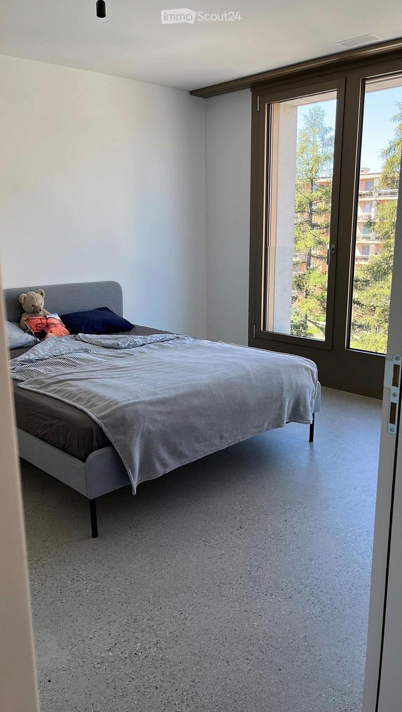 Modern bedroom with a grey bed, pillows, a blanket, and a teddy bear on the bed. Large sliding glass doors lead to a garden view.