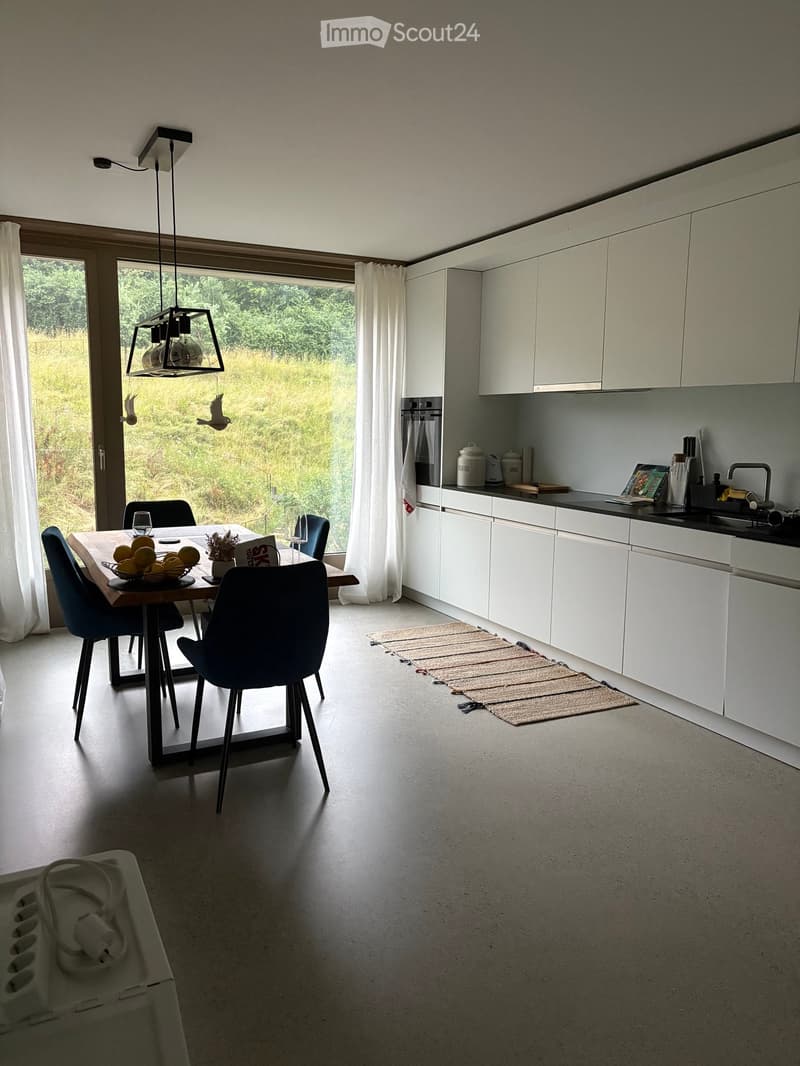Modern kitchen with white cabinets, a dining table with chairs, a hanging lamp, and a view of greenery through large windows.