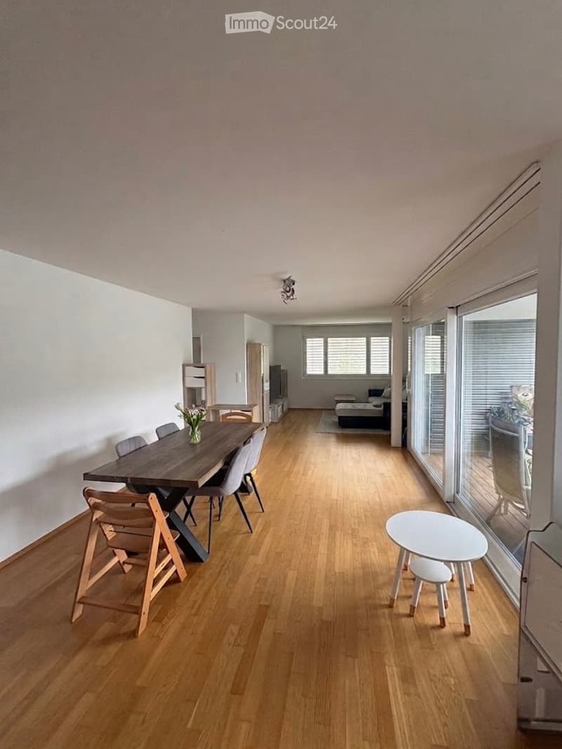 Open-plan living and dining area with wooden flooring, large windows, dining table with chairs, small round table with stools, shelf, TV stand, and sofa visible in the background.