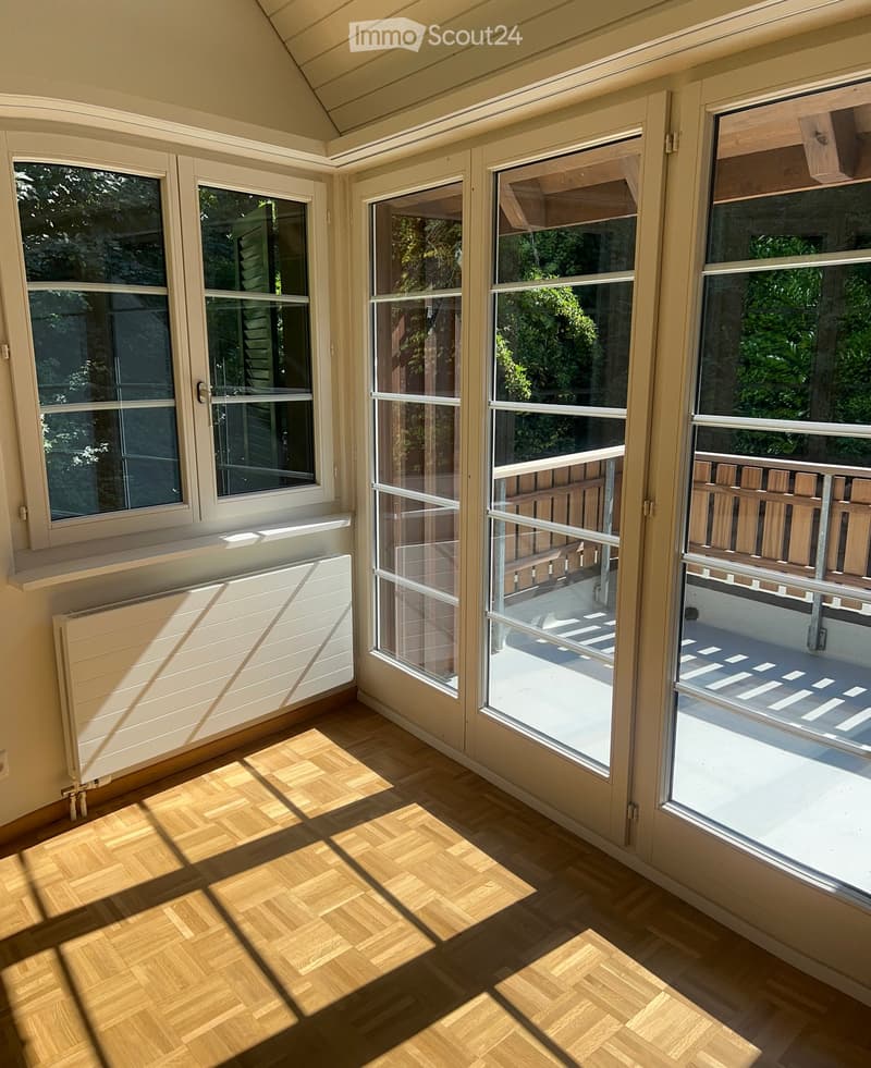 Bright sunroom with large windows and sliding glass doors leading to a balcony, wooden herringbone flooring, and a white radiator.