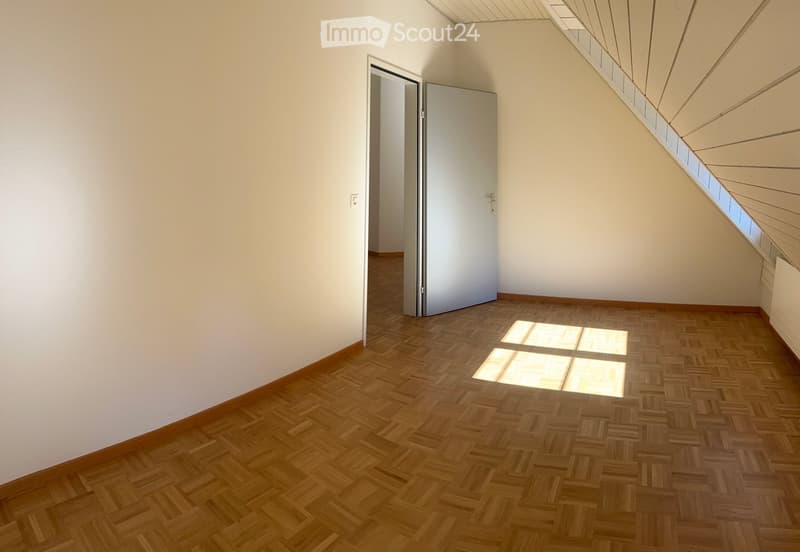 Empty room with wooden herringbone flooring, white walls, and a door leading to another space. The ceiling has skylights allowing natural light to enter.