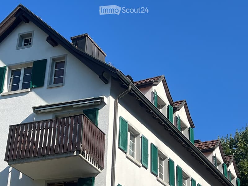 Two-story white building with green shutters, multiple windows, and a wooden balcony on the second floor.