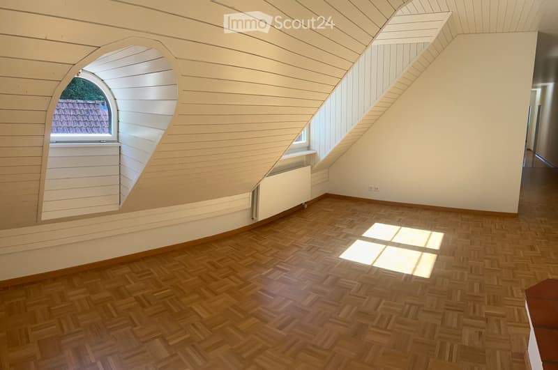 Empty attic room with wooden herringbone flooring, slanted wooden ceiling, and a large arched window offering a view of the roof.