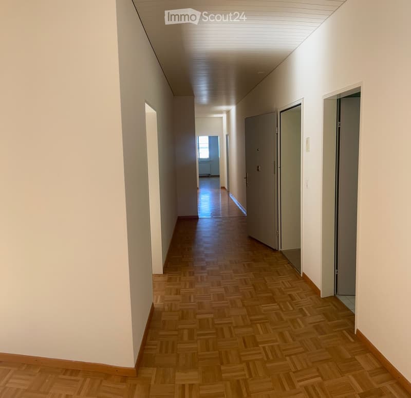 A long, narrow hallway with wooden herringbone flooring, white walls, and several closed doors on the right side. The ceiling has a ribbed design and recessed lighting.