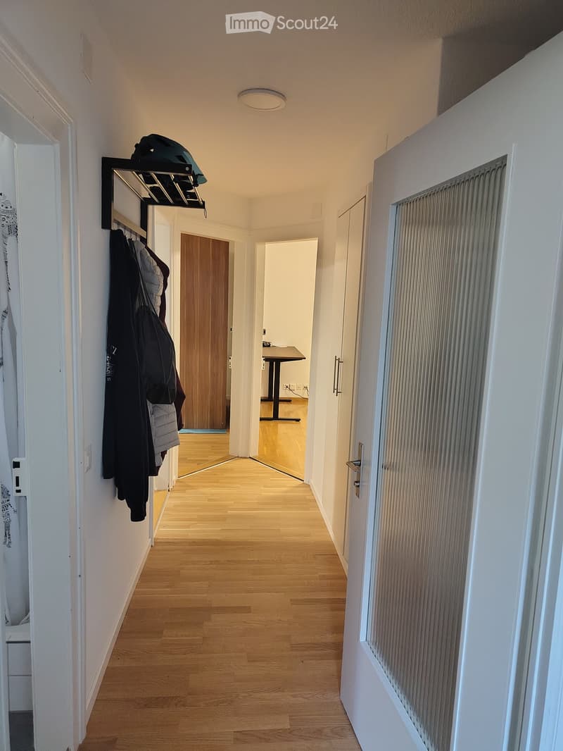 A narrow hallway with wooden flooring, white walls, and a coat rack holding jackets and bags. A door leads to another room with a small table visible in the background.