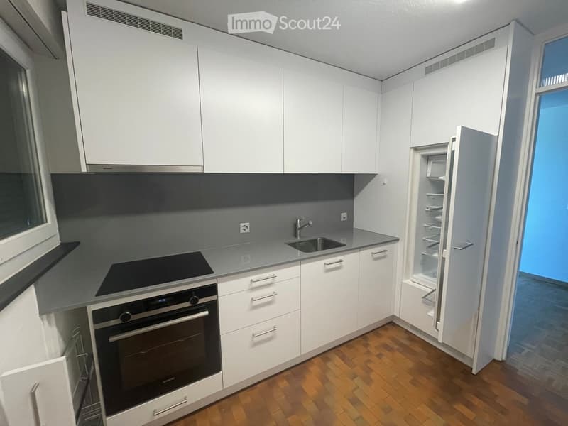 Modern kitchen with white cabinets, grey countertops, and wooden flooring. Features include a refrigerator, oven, sink, and faucet.