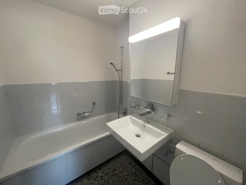 Modern bathroom with a bathtub, sink, toilet, and a large mirror with built-in lighting. The walls are tiled in a light grey color, and the floor has a patterned tile design.
