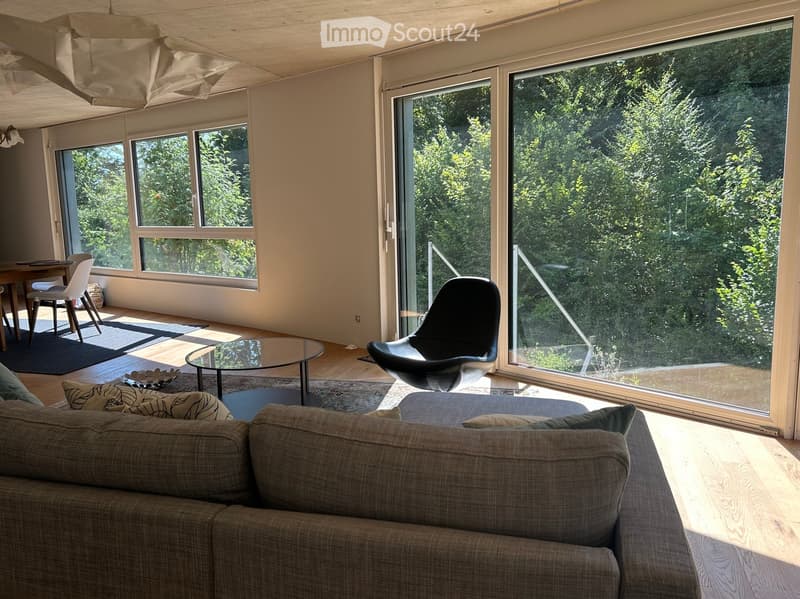 Bright living room with large windows and a view of greenery, featuring a sofa, coffee table, dining area, and modern chairs.