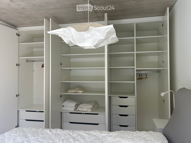 Modern bedroom with a neatly made bed, large white wardrobe with shelves and drawers, and a wall-mounted lamp.