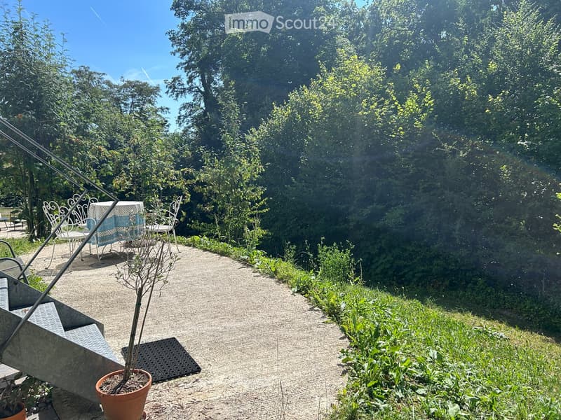 Garden path lined with greenery, small seating area with white metal chairs and a table, potted plant, and a concrete bench on the left side.