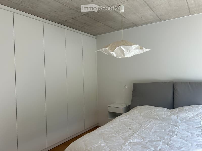 Modern bedroom with a white bed, grey pillows, a nightstand, and a large white wardrobe. The room features a contemporary design with a pendant light and wooden flooring.