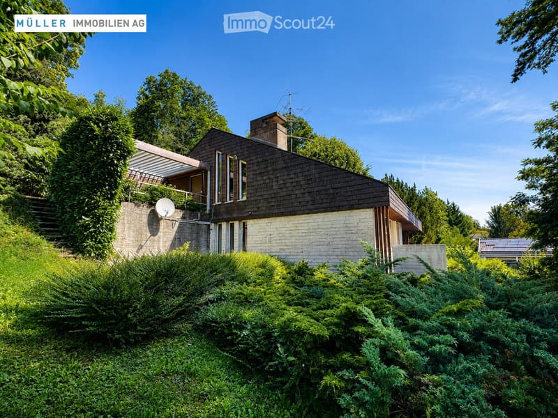 Modern two-story house with dark wooden roof, white siding, and large windows, surrounded by lush greenery and trees.