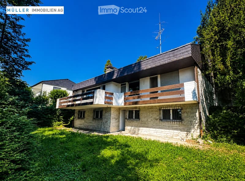 Two-story house with a raised ground floor, balcony, and modern design, surrounded by greenery.