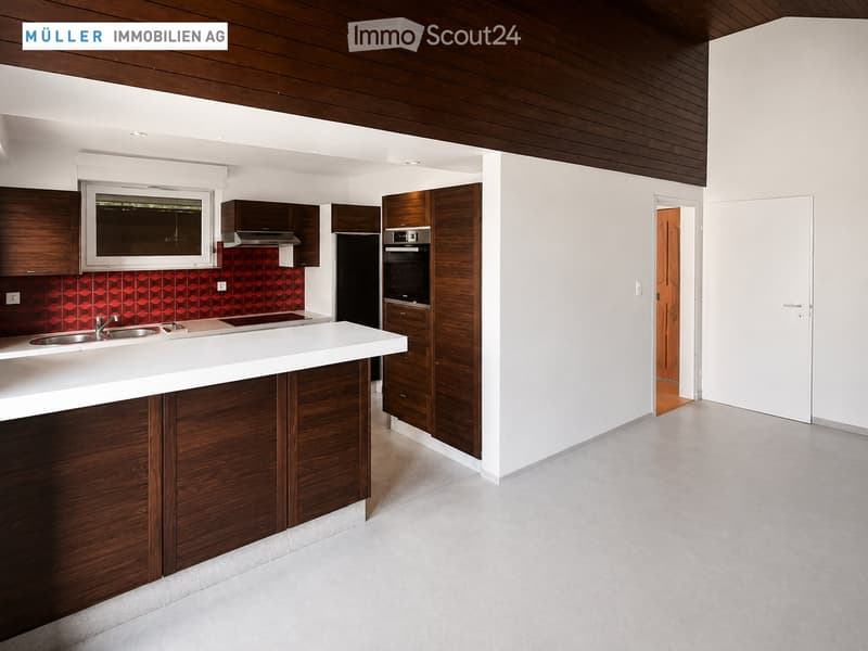 Modern kitchen with white countertops, wooden cabinets, stainless steel sink, and built-in oven. Features a red backsplash and large window.