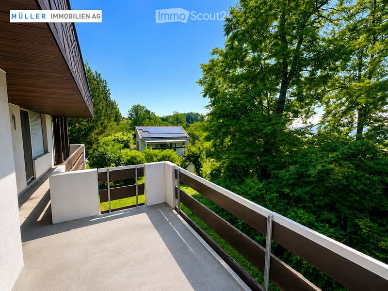 Balcony with wooden railing, overlooking a lush green garden and a neighboring house with solar panels on the roof.