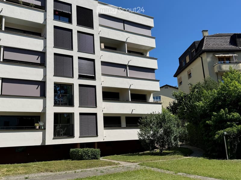 Modern multi-story apartment building with balconies, large windows, and greenery in front.