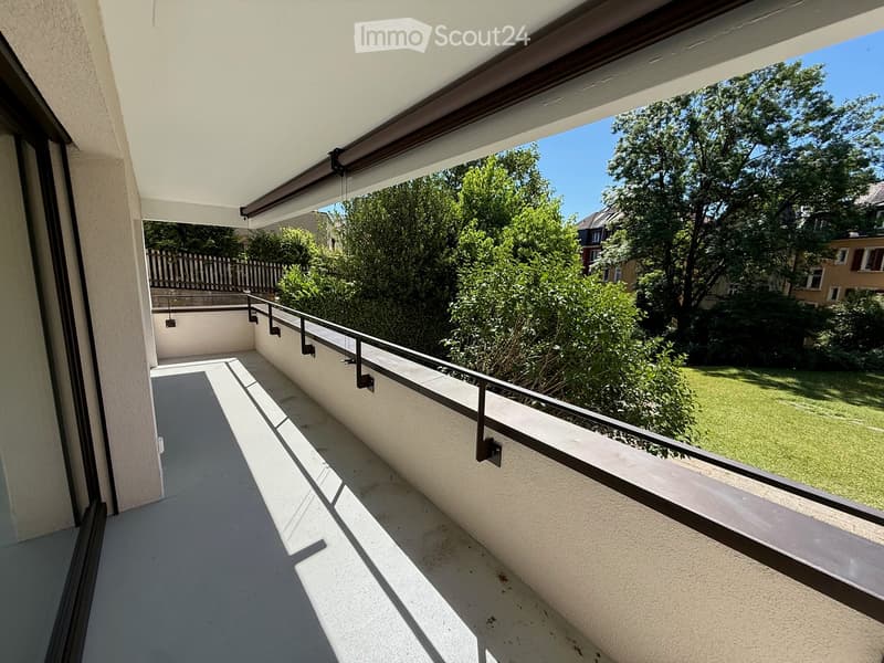 Balcony with modern design, white walls, metal railing, and a retractable awning, overlooking a green garden area.