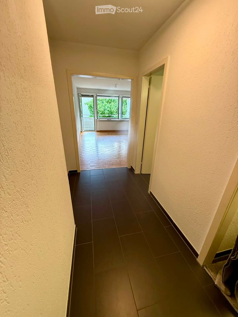 A narrow hallway with dark tiled flooring and light-colored walls, leading to an open area with large windows and wooden flooring.