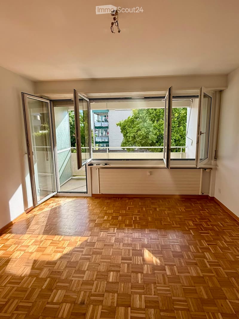 Empty room with wooden herringbone flooring, large windows with sliding glass doors, radiator, and visible electrical outlet.