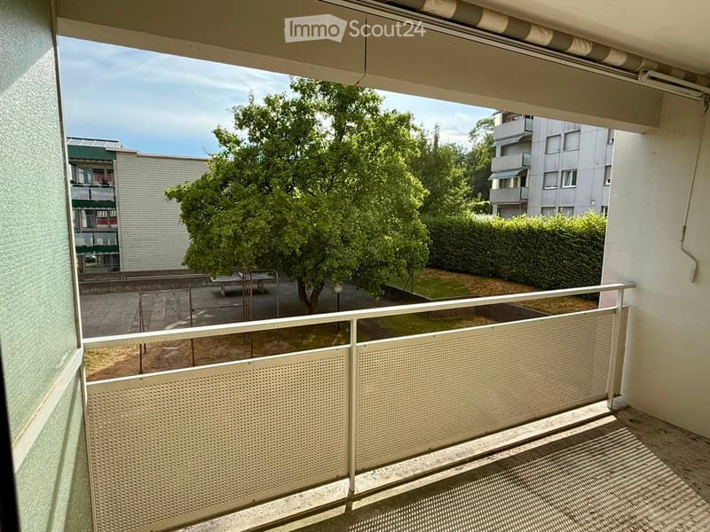 Balcony with metal railing, overlooking a courtyard with trees, benches, and neighboring buildings.