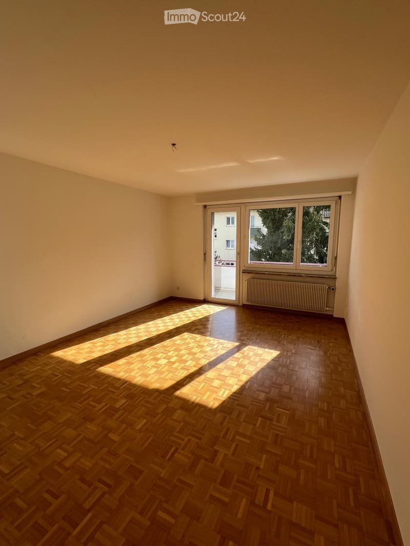 Empty room with wooden parquet flooring, white walls, and large windows with blinds. Natural light streams in, casting shadows on the floor.