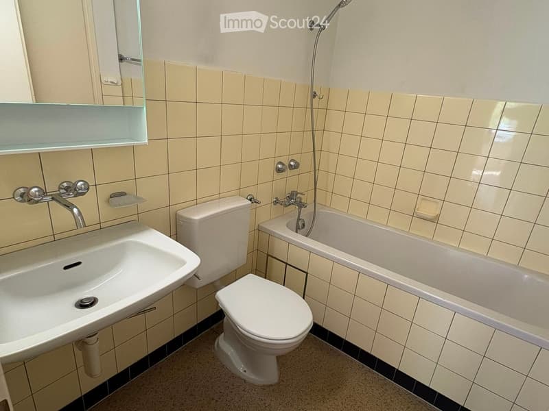 Bathroom with beige tiled walls, a white sink, toilet, bathtub, and a glass shelf above the sink.