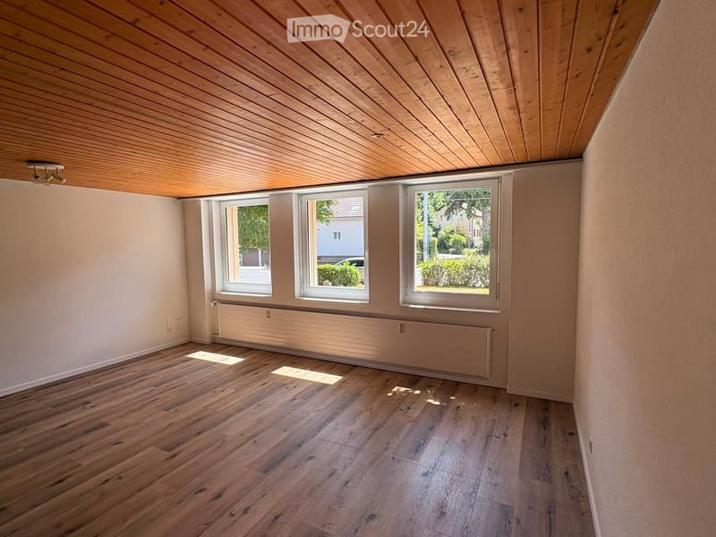 Empty room with wooden flooring, light-colored walls, and large windows letting in natural light. The ceiling is wooden with recessed lighting.