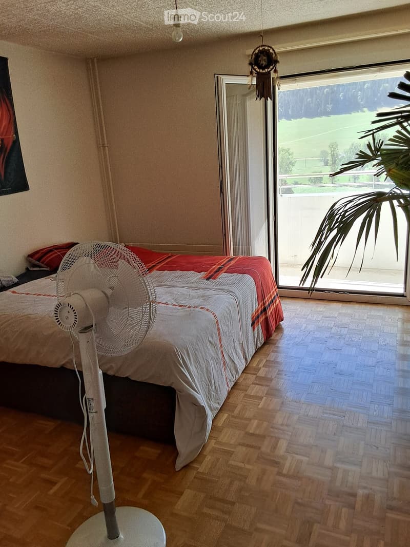 Bedroom with wooden flooring, a large bed with red and white bedding, a standing fan, and a large window with a view of a green field.