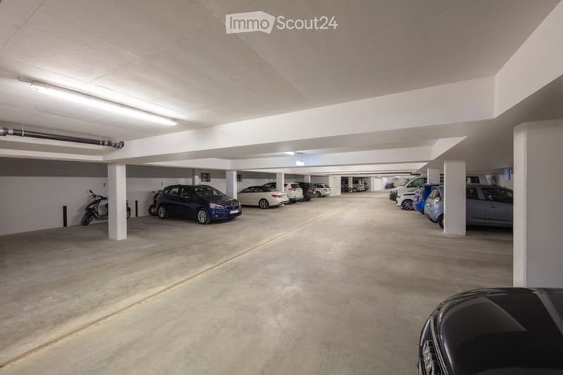 Underground parking area with several parked cars, including sedans, SUVs, and a scooter, under a concrete ceiling with fluorescent lighting.