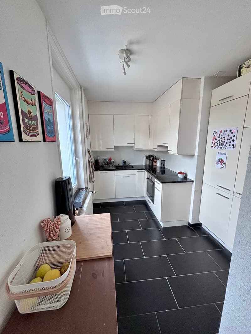 Modern kitchen with white cabinets, black countertops, and stainless steel appliances. Features a refrigerator, oven, sink, and a small kitchen island with a basket of lemons.