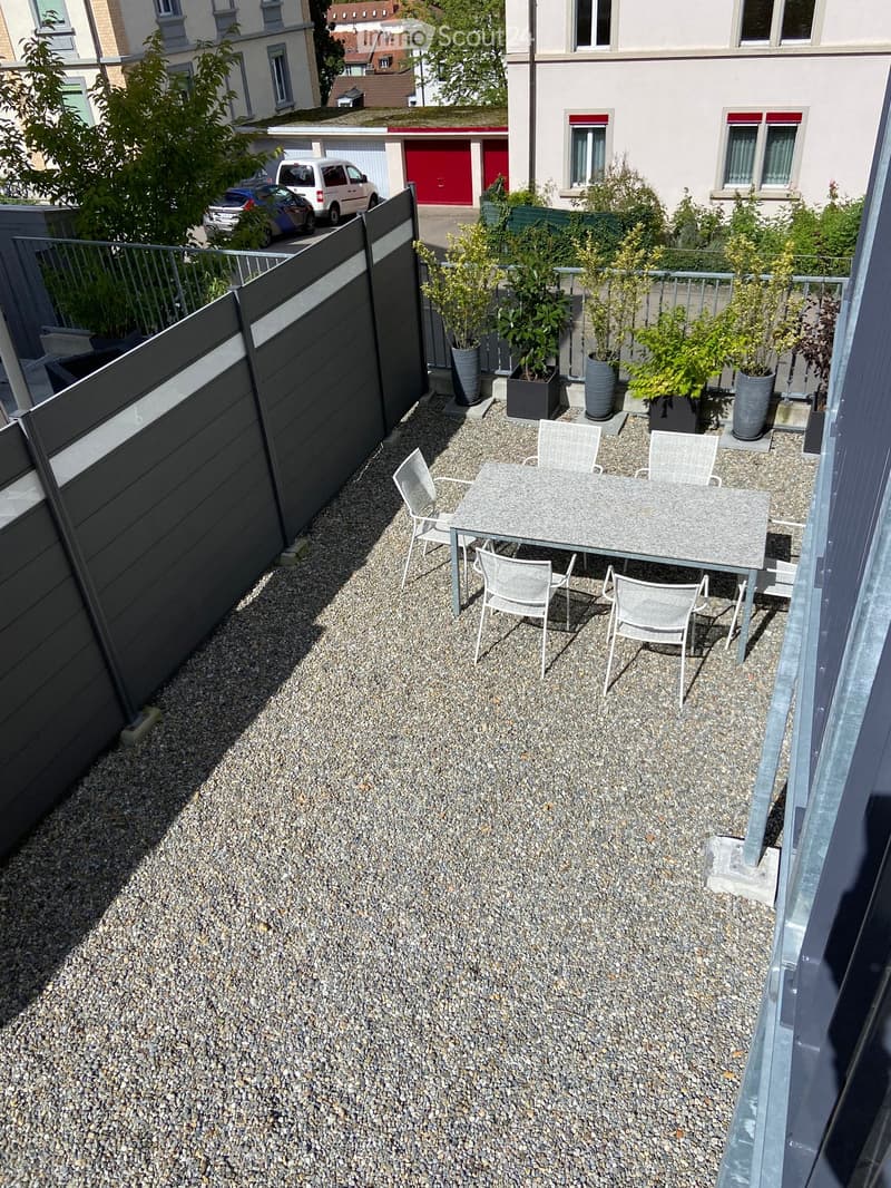 Compact outdoor patio with a gravel surface, a small table with four chairs, and several potted plants. The area is enclosed by a black fence and is adjacent to a residential building.