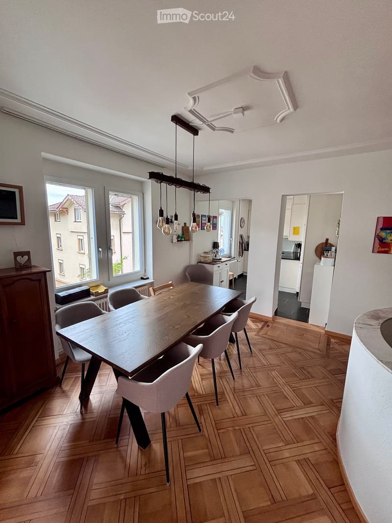 Dining area with a wooden dining table, six chairs, a kitchen area with a counter, and a refrigerator. The room has wooden flooring and large windows with a view of the street.