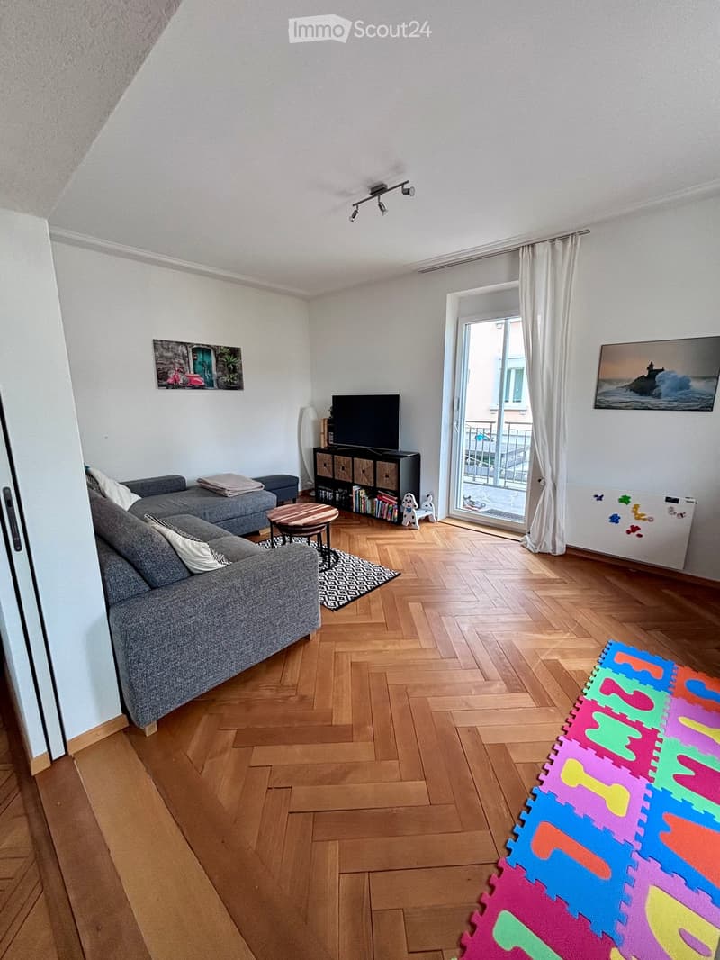 Living room with wooden flooring, grey sofa, coffee table, TV stand, bookshelf, and a small balcony visible through large windows.