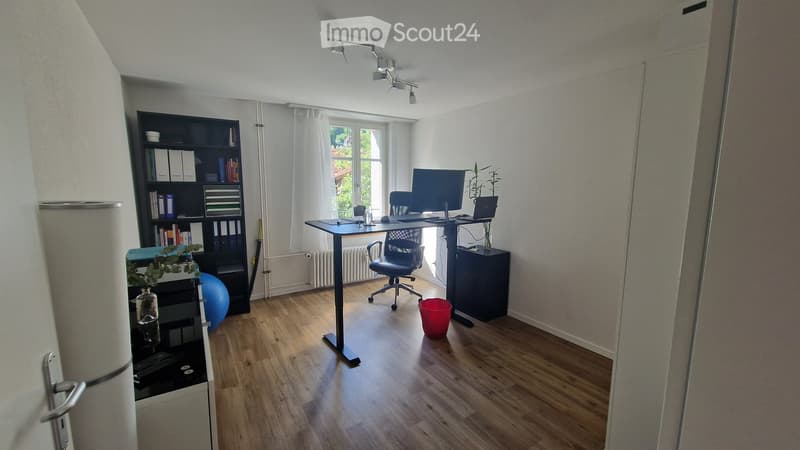 A modern, well-lit home office with a wooden floor, large window with white curtains, a standing desk with a laptop and screen, a black office chair, a bookshelf filled with books and binders, a blue exercise ball, and a small plant on a side table.