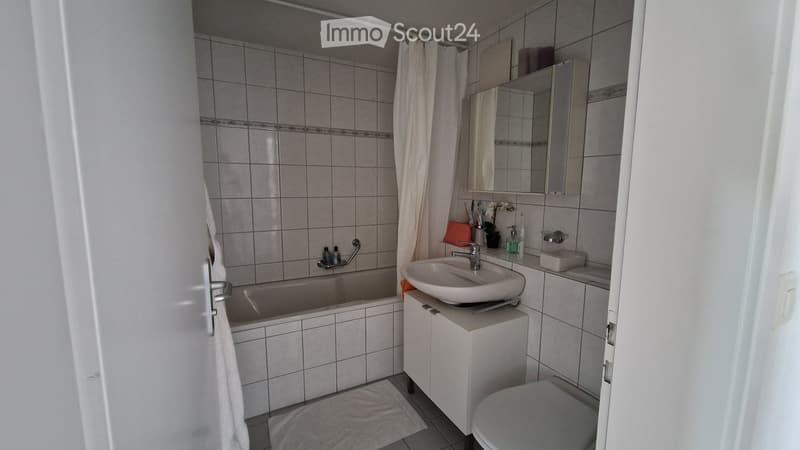 Bathroom with a bathtub, sink, toilet, mirror, and cabinet. The walls are tiled, and there is a shower curtain in the background.