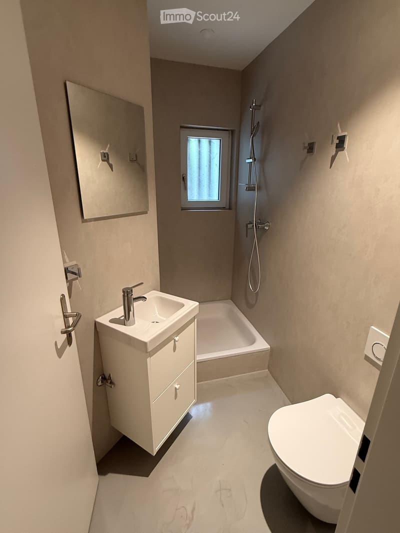 Modern bathroom with a white sink, bathtub, toilet, mirror, and shower. Features a small window and sleek, contemporary design.