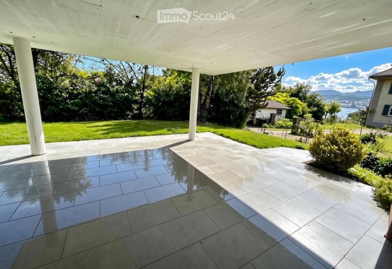 Covered outdoor patio with polished tile flooring, white columns, and a scenic view of a green lawn, trees, and distant water.