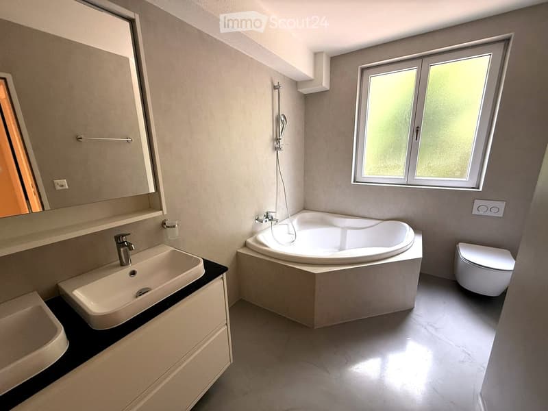 Modern bathroom with a white bathtub, sink, toilet, and large mirror. Features a window with frosted glass and sleek, contemporary design.