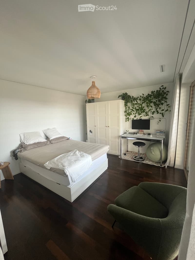 Modern bedroom with a white bed, grey bedding, a green armchair, a desk with a computer, a hanging lamp, and a large plant in the corner.
