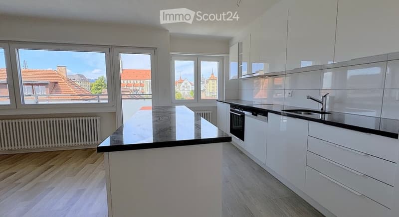 Modern kitchen with white cabinets, black countertop, stainless steel sink and faucet, and a central island. Large windows offer a view of the cityscape.