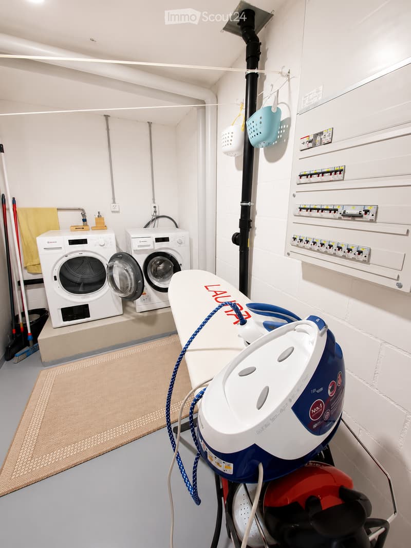 A compact utility room with a washing machine, dishwasher, ironing board, and cleaning supplies.