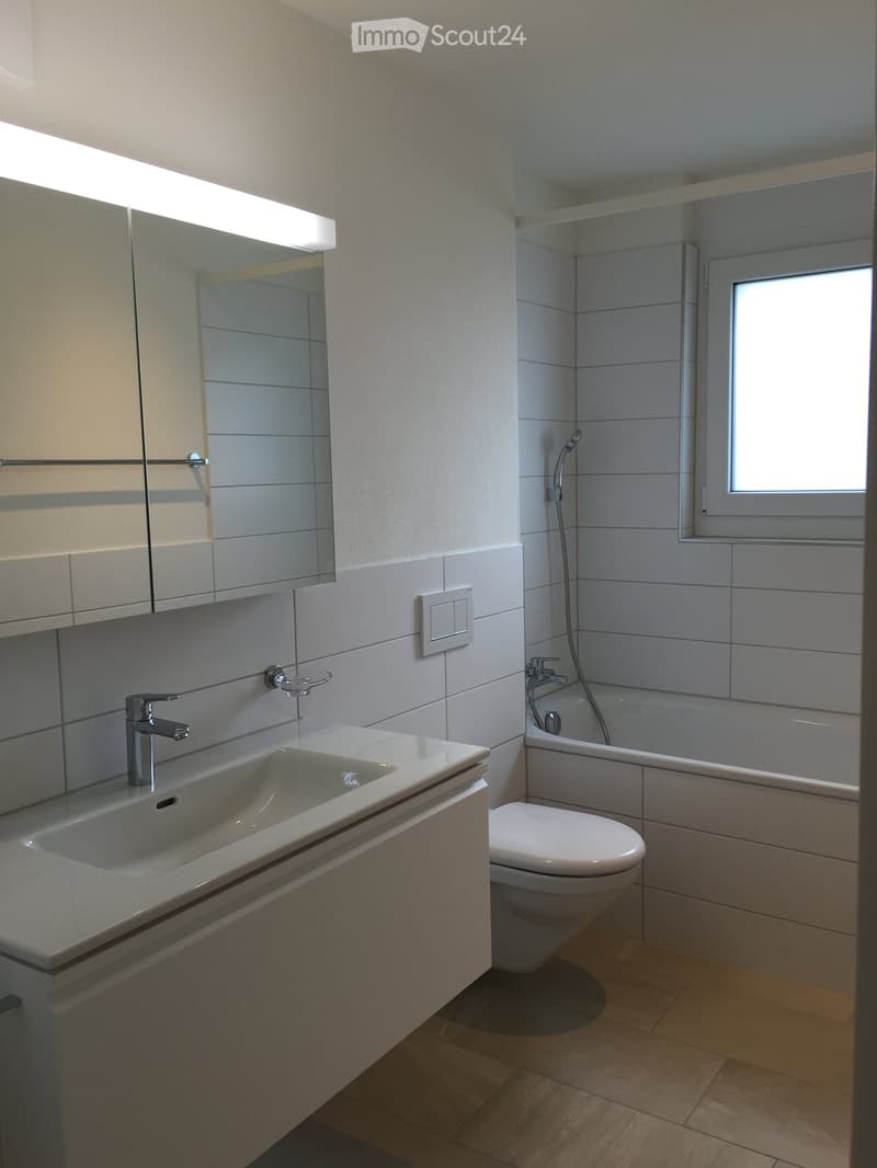 Modern bathroom with a white sink, mirror, toilet, bathtub, and showerhead. Light-colored tiles and wooden flooring.