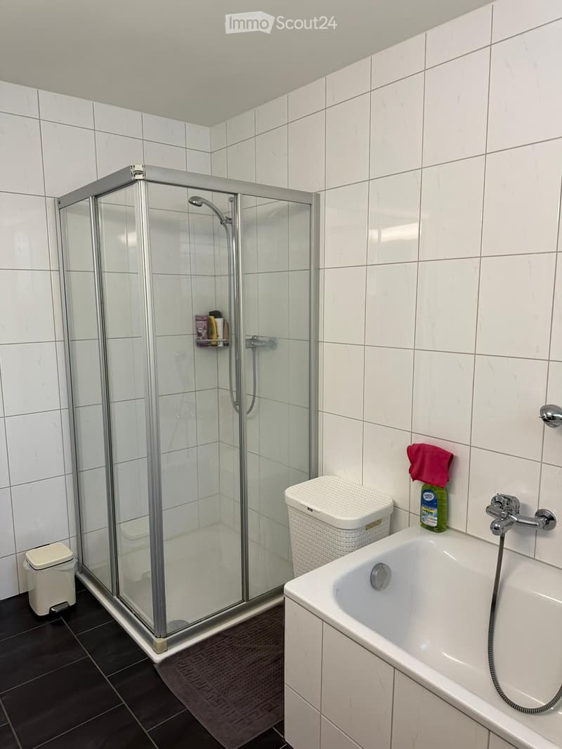 Modern bathroom with white tiled walls, a bathtub, a glass-enclosed shower, a white trash bin, and a cleaning bottle on the edge of the tub.