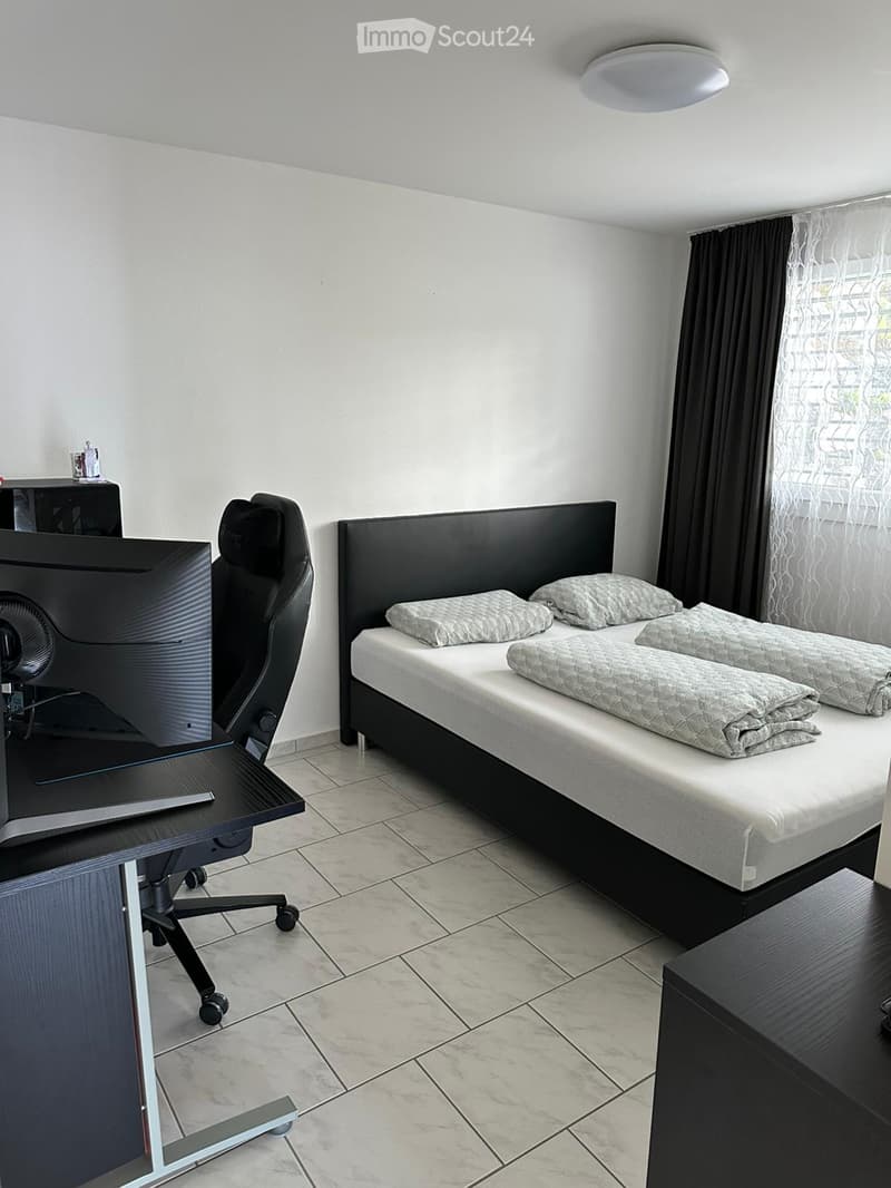 A modern bedroom with a double bed, a desk with a chair, and a large screen. The room features tiled flooring and large windows with curtains.