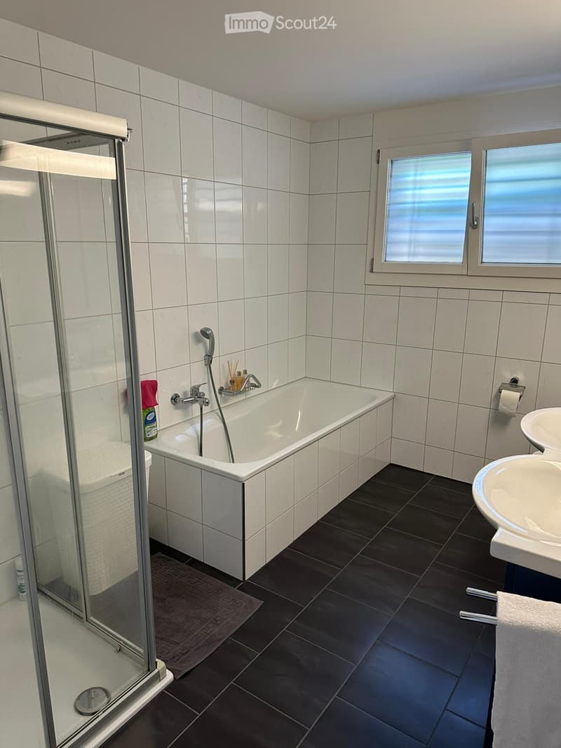 Modern bathroom with white tiles, a bathtub, a glass-enclosed shower, a sink, and a window with blinds.