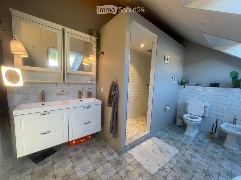 Modern bathroom with a double sink, large mirror, shower, toilet, and bidet. Features white cabinets, grey tiles, and decorative lighting.