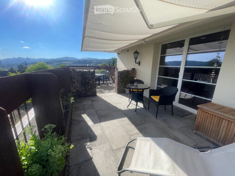 Balcony with a view, featuring a table, chairs, and a lounge chair, with a covered awning and a scenic mountain backdrop.